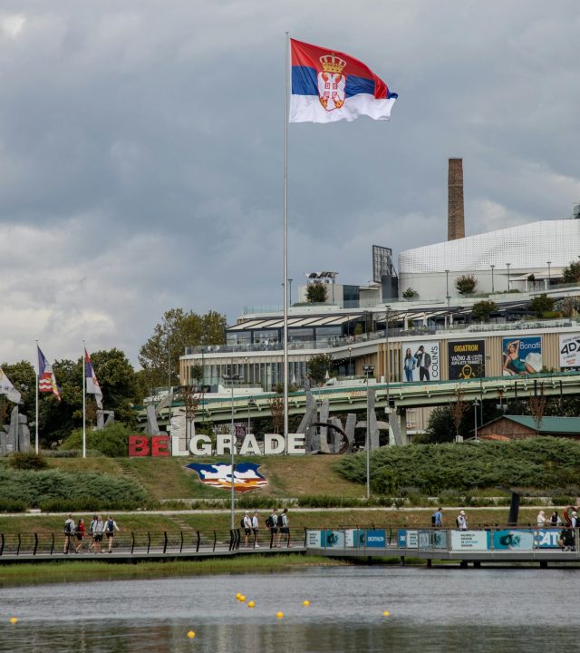 Serbian flag flies high over Belgrade's waterfront with cityscape and park view.